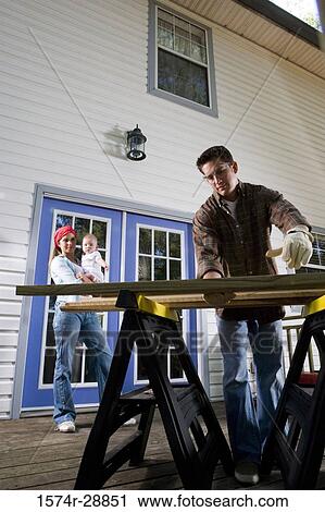 Stock Image - Young woman carrying baby while young man holding wood plank. Fotosearch