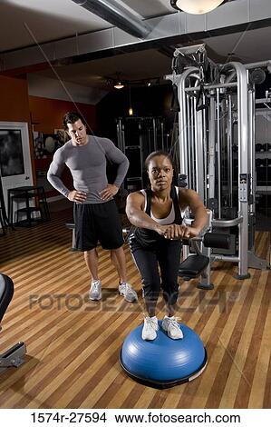 Young woman exercising on a fitness ball while trainer looking her in the gym View Large Photo Image Picture - Young woman exercising on a fitness ball while trainer looking her in the gym. Fotosearch