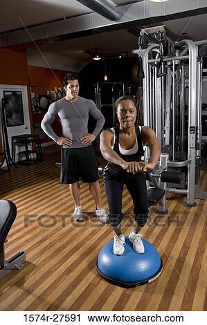 Young woman exercising on a fitness ball while trainer looking her in the gym View Large Photo Image Stock Image - Young woman exercising on a fitness ball while trainer looking her in the gym. Fotosearch