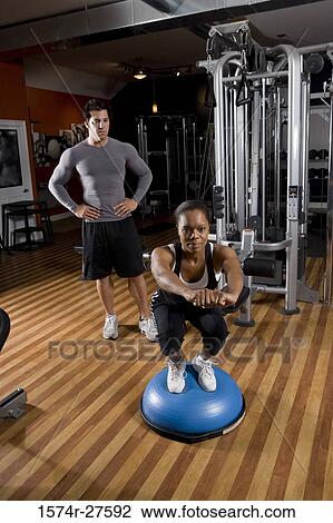 Young woman exercising on a fitness ball while trainer looking her in the gym View Large Photo Image Stock Image - Young woman exercising on a fitness ball while trainer looking her in the gym. Fotosearch