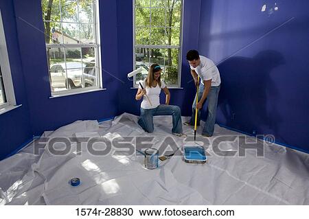Stock Image - Young woman holding paint roller while young man preparing color. Fotosearch