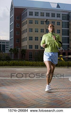 Picture - Young woman in sports clothing jogging on walkway with building in the background. Fotosearch