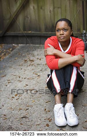 Stock Image - Young woman in sports clothing sitting in front of a wooden door. Fotosearch