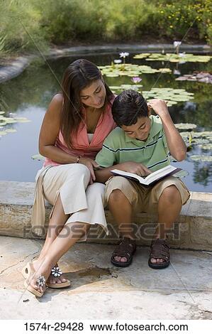 Stock Photo - Young woman sitting with her son by pond reading book. Fotosearch