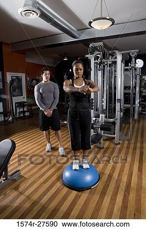 Young woman standing on a fitness ball while trainer in the background in the gym View Large Photo Image Stock Image - Young woman standing on a fitness ball while trainer in the background in the gym. Fotosearch