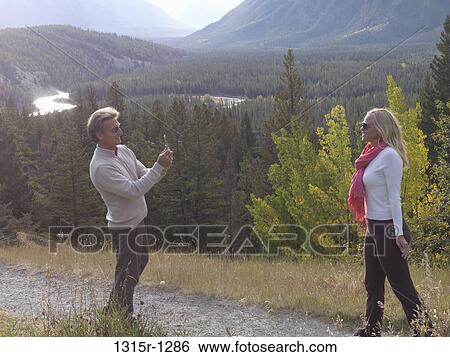 Stock Photograph - Canada, Alberta, couple taking pictures with cell phone on path through mountain meadow,. Fotosearch