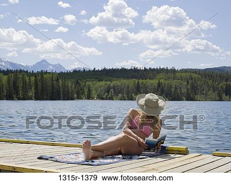 Stock Photo - Canada, Alberta, Jasper National Park, woman sitting on mountain lake dock and reading. Fotosearch