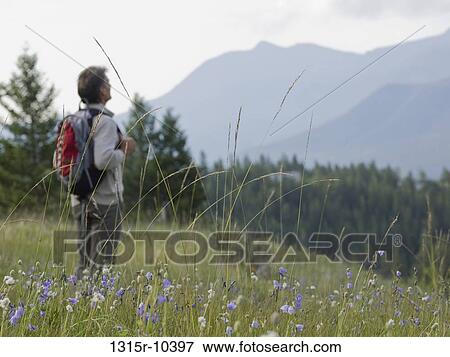 Stock Photo - Canada, Alberta, Rear view of man with knapsack standing in mountain meadow, looking at mountains. Fotosearch