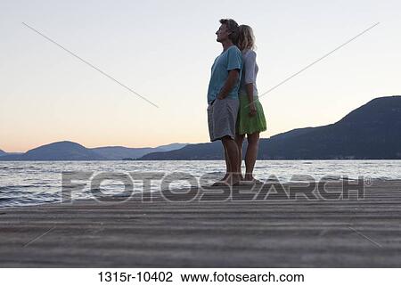 Stock Image - Canada, British Columbia, Okanagan Lake, Couple standing back to back on lakeside wharf. Fotosearch