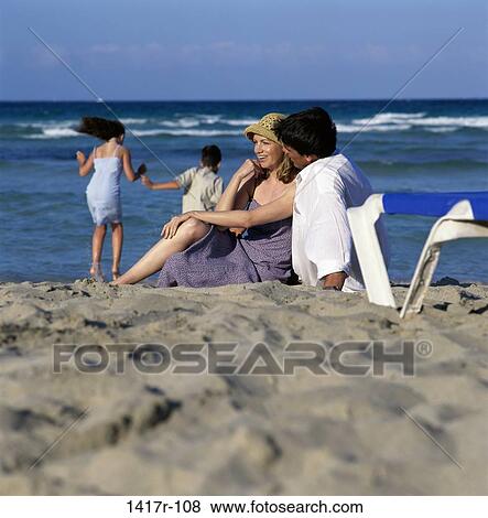 Stock Photo - Father and mother at the beach with their daughter and son playing in the background. Fotosearch