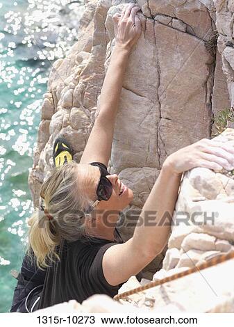 Stock Image - Italy, Liguia, Female climber ascends steep rock above shimmering sea. Fotosearch
