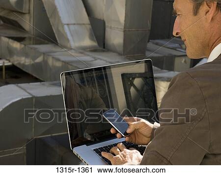 Stock Image - Italy, Piedmont, Businessman working on cell phone and laptop. Fotosearch