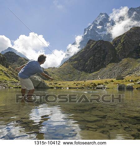Stock Image - Italy, Piedmont, Hiker skipping stones across mountain lake. Fotosearch