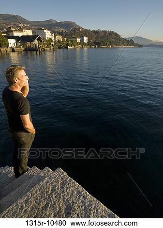 Stock Image - Italy, Piedmont, Lake Maggiore, Man talking on cell phone at lake edge. Fotosearch