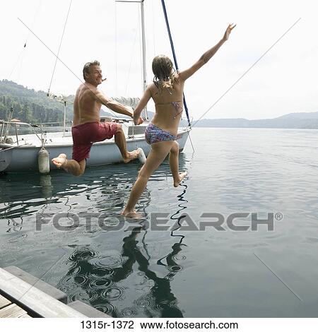 Stock Image - Italy, Piedmont, Lake Orta, Father and daughter jump into water from dock. Fotosearch