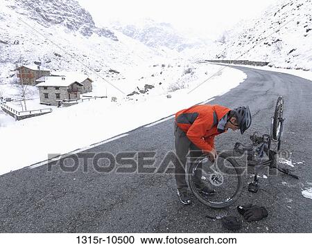 Stock Image - Italy, Piedmont, Mountain biker fixing bike on snowy mountain road. Fotosearch