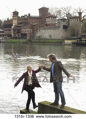 Stock Photo - Italy, Piedmont, Turin, man helping woman to climb wall by river. Fotosearch