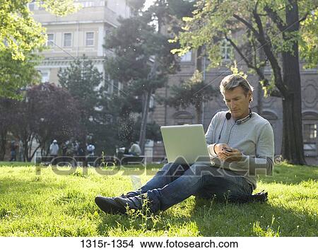 Italy, Piedmont, Turin, Man listening to MP3 player while working on laptop in park View Large Photo Image Picture - Italy, Piedmont, Turin, Man listening to MP3 player while working on laptop in park. Fotosearch