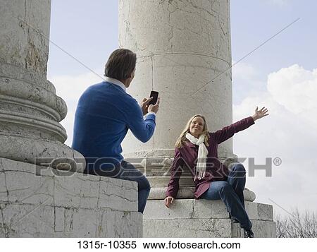 Stock Photography - Italy, Piedmont, Turin, Man photographing woman sitting on marble pillar with camera phone. Fotosearch