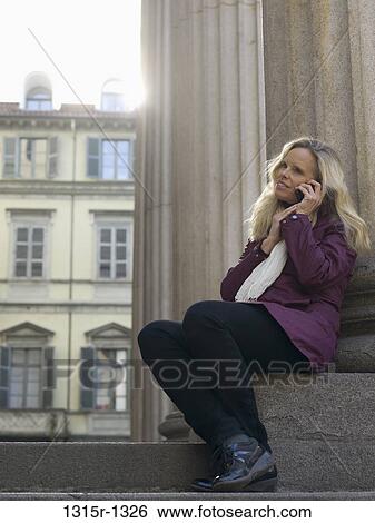 Stock Photograph - Italy, Piedmont, Turin, Woman talking on cell phone while sitting on step below pillars. Fotosearch