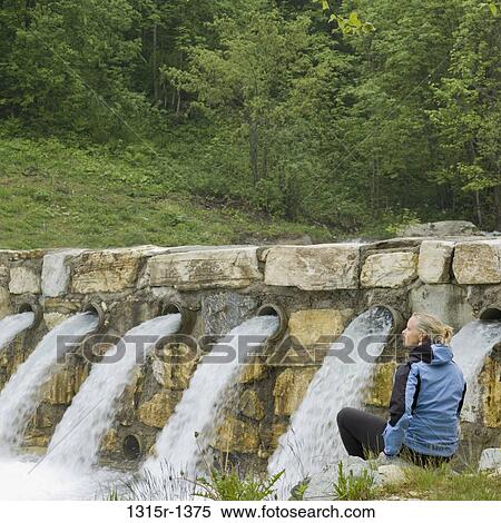 Stock Photography - Italy, Piedmont, woman sitting on rock in front of creek waterspouts. Fotosearch