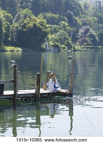 Stock Photography - Italy, Piedmont, Woman sitting on wooden pier, splashing feet in Lake Orta. Fotosearch