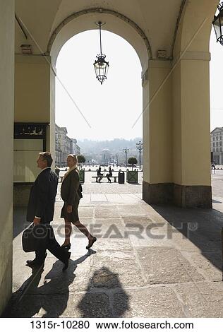 Stock Image - Italy, Turin, Business couple walk through porticos in front of piazza. Fotosearch
