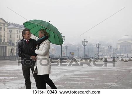 Stock Image - Italy, Turin, Couple walking through plaza under umbrella. Fotosearch
