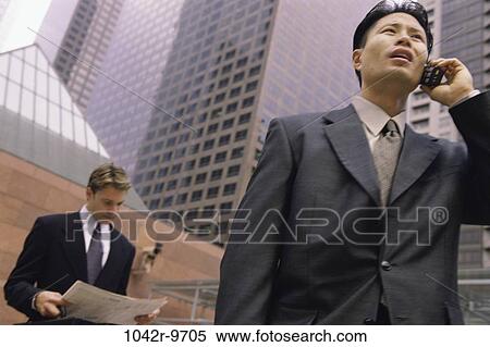 Stock Photography - Low angle view of a businessman talking on a mobile phone and reading a newspaper. Fotosearch