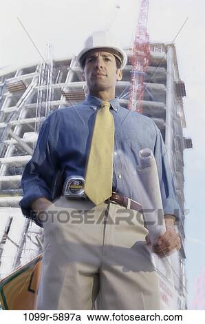 Stock Image - Low angle view of an engineer at a construction site holding blueprints. Fotosearch