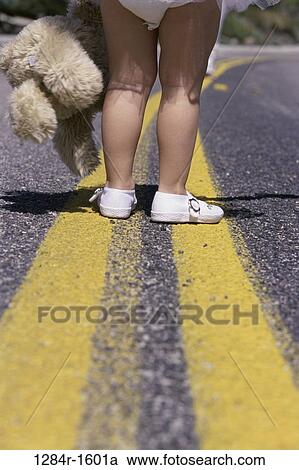 Stock Image - Low section view of a girl's feet standing on a road holding a stuffed toy. Fotosearch