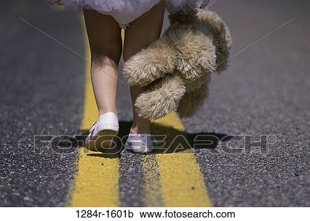 Stock Image - Low section view of a girl's feet walking on a road holding a stuffed toy. Fotosearch