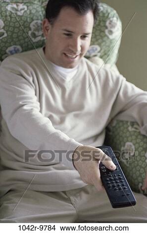 Picture - Mid adult man sitting on a couch holding a remote control. Fotosearch