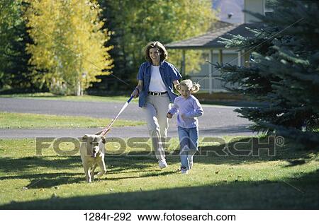 Stock Image - Mother and daughter walking their pet dog on a lawn. Fotosearch