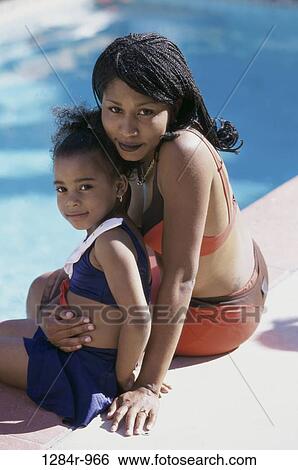 Stock Photograph - Mother holding her daughter sitting at the edge of a swimming pool. Fotosearch