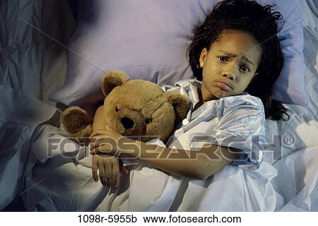 Stock Image - Portrait of a girl lying in bed holding a teddy bear. Fotosearch