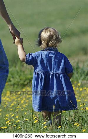Stock Image - Rear view of a baby girl walking in a field holding her mother's hand. Fotosearch