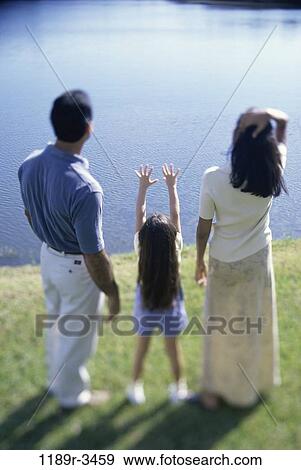 Stock Photo - Rear view of a mother and father with their daughter standing in front of a lake. Fotosearch