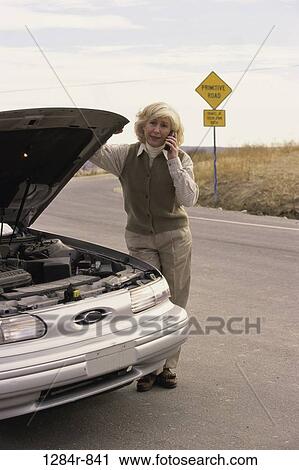 Stock Image - Senior woman standing near a broken down car talking on a mobile phone. Fotosearch