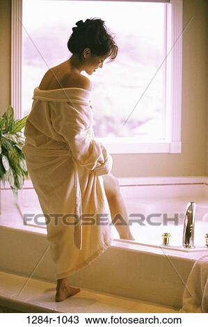 Stock Image - Woman in a bath robe standing at the edge of a bathtub. Fotosearch