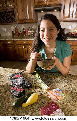 Picture - Asian girl in kitchen holding bowl and chopsticks. Fotosearch