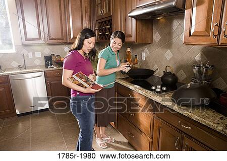 Stock Image - Asian mother and daughter cooking in wok looking at cookbook. Fotosearch