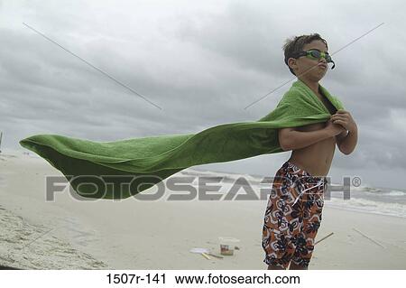 Stock Image - Boy holding a towel standing at the beach. Fotosearch