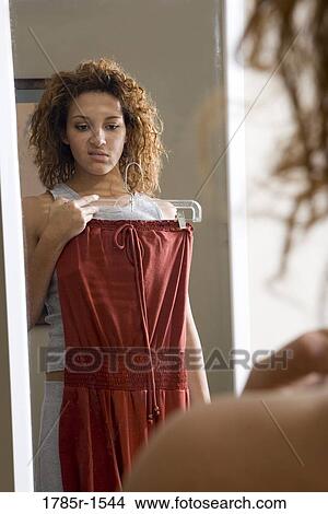 Close-up of a young woman standing in front of a mirror, frowning while inspecting a dress on a hanger View Large Photo Image Picture - Close-up of a young woman standing in front of a mirror, frowning while inspecting a dress on a hanger. Fotosearch