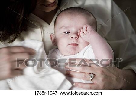 Stock Photo - Close-up of an infant rubbing his eyes while being held by his mother. Fotosearch