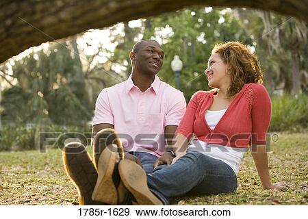 Close-up of smiling young multi-ethnic couple sitting on grass outdoors View Large Photo Image Stock Photo - Close-up of smiling young multi-ethnic couple sitting on grass outdoors. Fotosearch