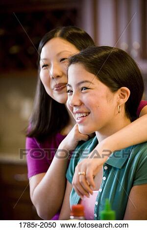 Stock Image - Close-up portrait of an Asian mother embracing her Asian-Caucasian daughter in the kitchen. Fotosearch