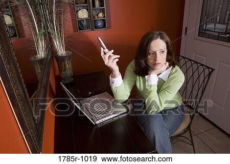 Stock Photo - Contemplative young woman holding a mobile phone while sitting at desk with laptop. Fotosearch