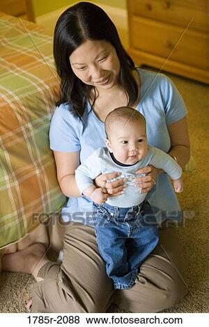 Stock Photo - Elevated view of an Asian mother sitting on the floor holding an infant boy on her lap. Fotosearch