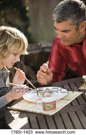 Stock Image - Father showing son how to eat sushi with chopsticks. Fotosearch